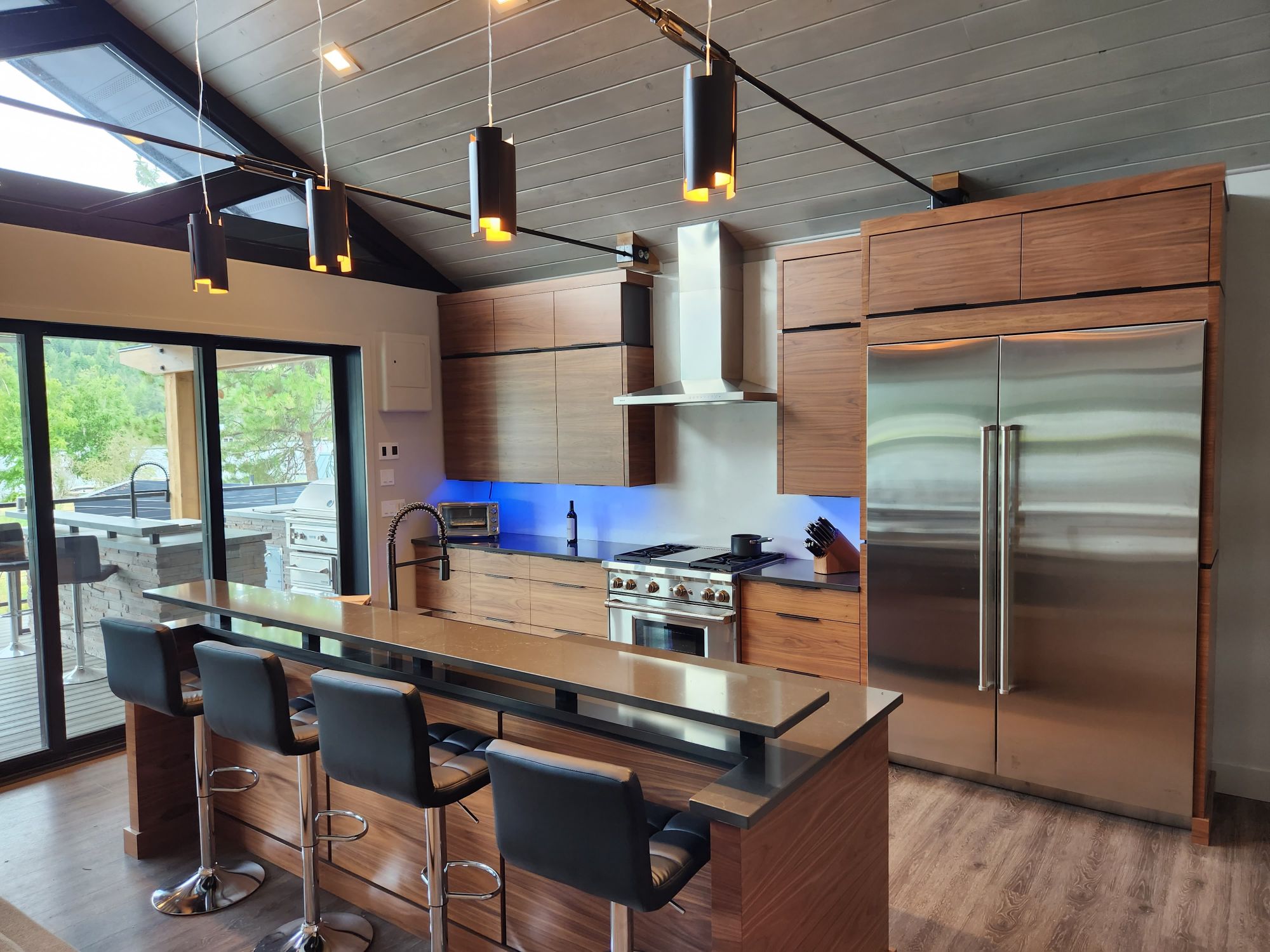 A kitchen with flat-panel wood cabinets, a two-tier island with black bar stools, a stainless range and refrigerator, pendant lights and a sloped plank ceiling with skylights