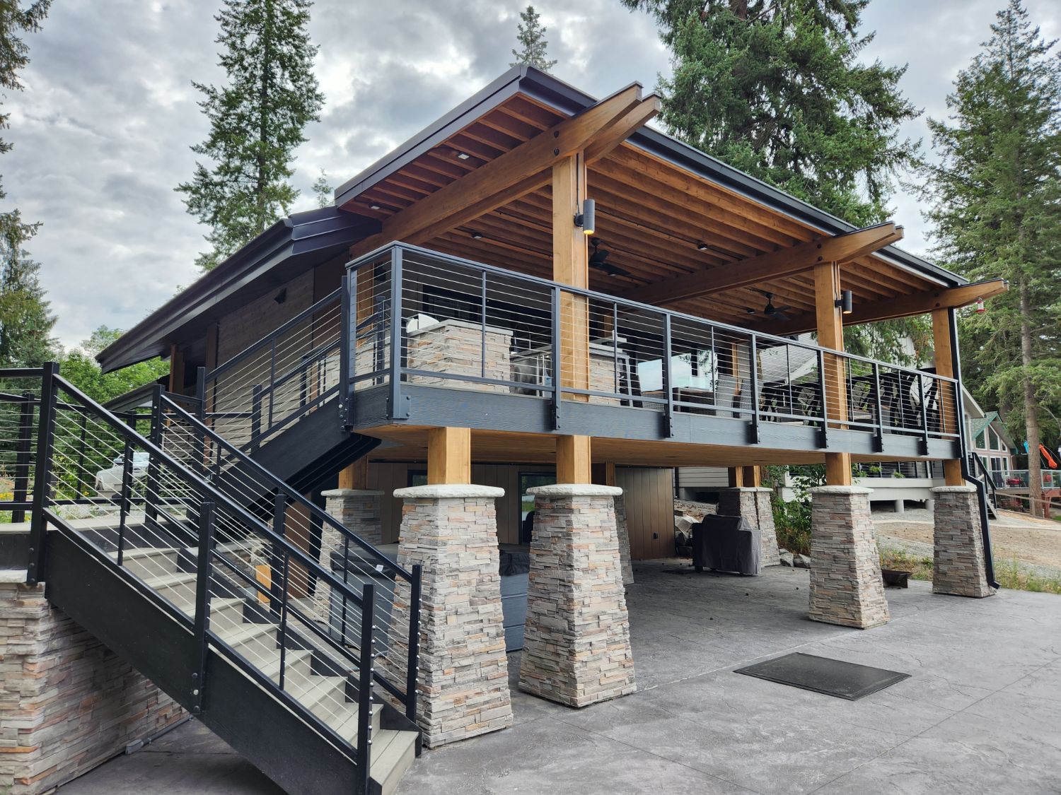 A home with a covered upper deck on heavy timber beams, black cable railings, an exterior stair and stacked-stone piers, among tall firs