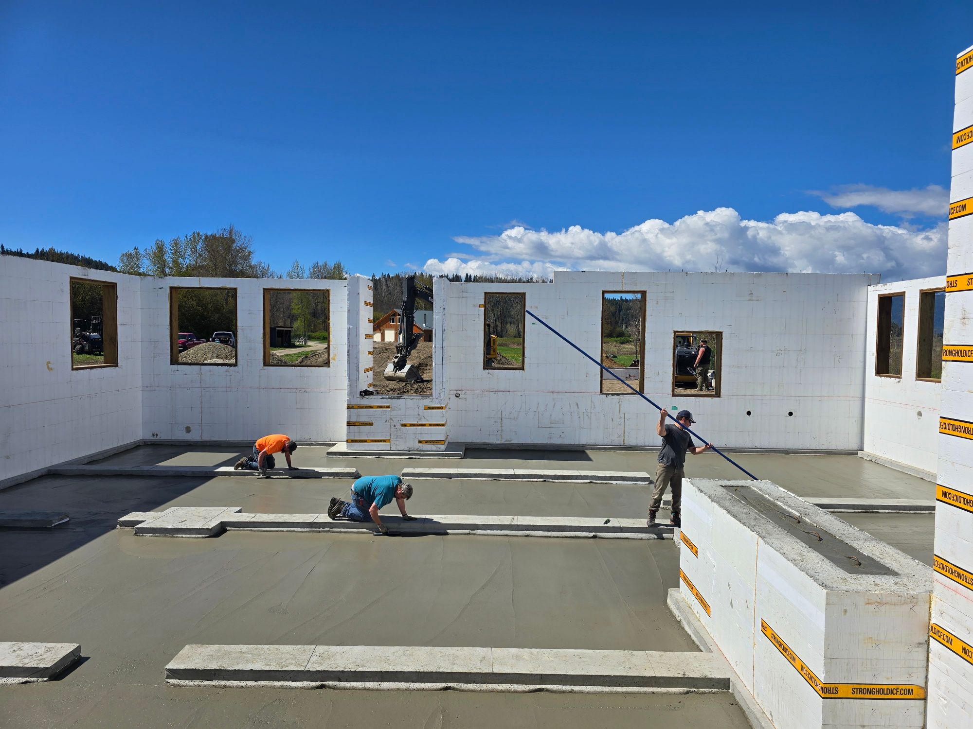 A crew finishing a fresh concrete slab inside insulated concrete form walls with open window bays, under a bright blue sky